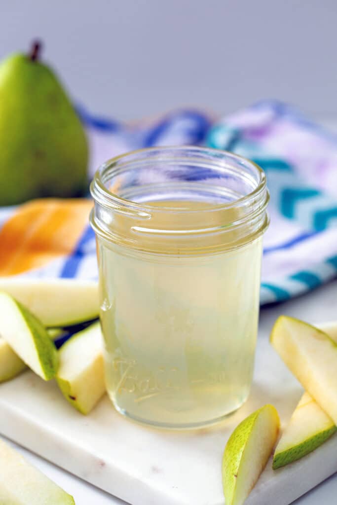 Overhead view of a small jar of pear simple syrup with sliced pears all around and whole pear in background.