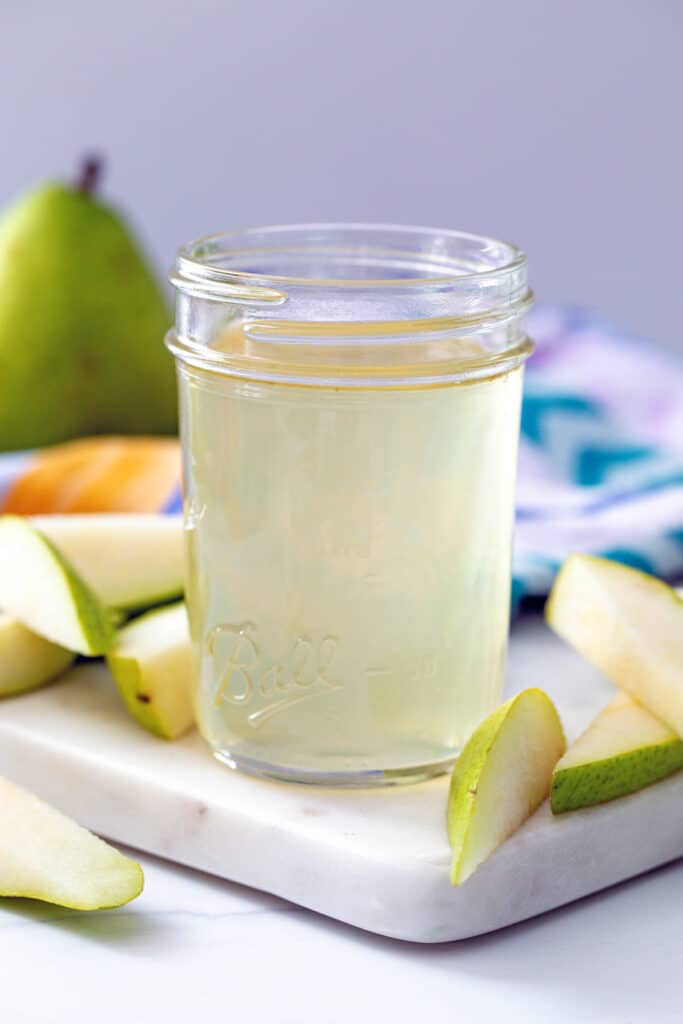 Head-on view of a small mason jar of pear simple syrup with sliced pears and a while pear in the background.