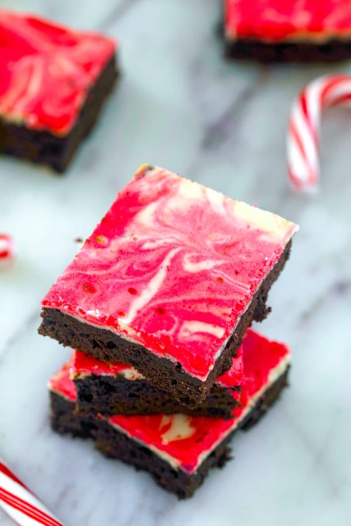 Overhead view of three peppermint cheesecake brownies stacked on each other on a marble surface with more brownies and candy canes in the background