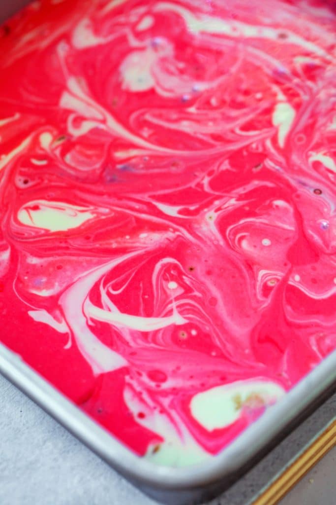 Overhead view of pan filled with brownie batter base and peppermint cheesecake topping with red and white colors swirled, ready to go into oven