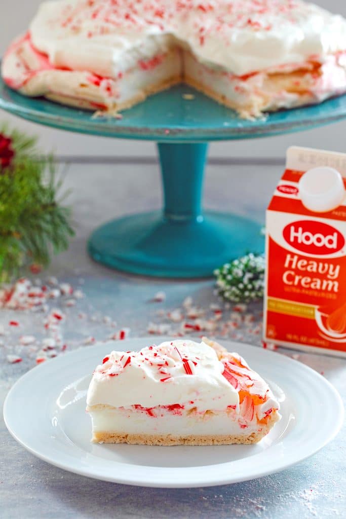 Head-on view of a slice of peppermint pavlova topped with white chocolate whipped cream on a white plate with a carton of Hood Cream and a cake stand with rest of pavlova in the background, surrounded by crushed candy canes