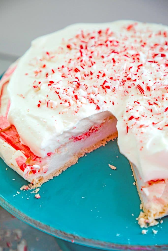 View of peppermint pavlova on cake stand topped with white chocolate whipped cream and candy cane crumbles with slice taken out