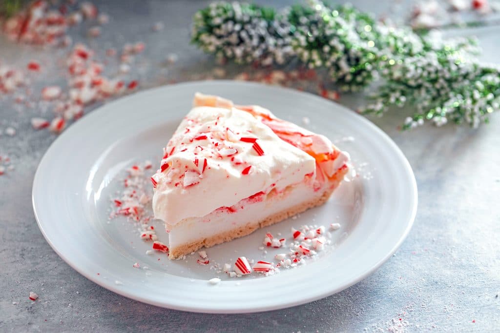 Landscape overhead close-up of a slice of peppermint pavlova on a white plate topped with white chocolate whipped cream and crushed candy canes, surrounded by crushed candy canes and green holly in the background