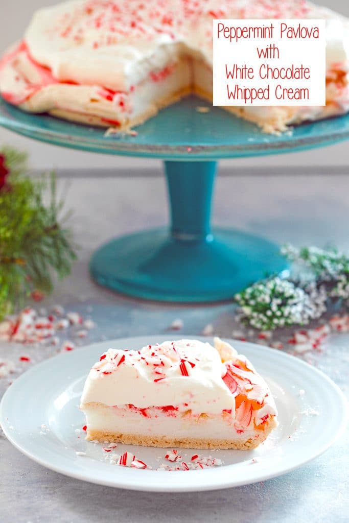 Head-on view of slice of peppermint pavlova with white chocolate whipped cream on a white plate with a teal cake stand with rest of pavlova in the background, surrounded by crushed candy canes with recipe title at top of image