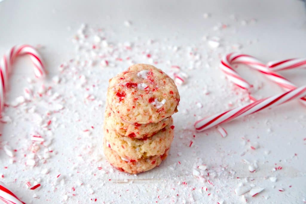 Landscape photo of four peppermint sugar cookies stacked on each other on white surface surrounded by crushed candy canes and whole candy canes