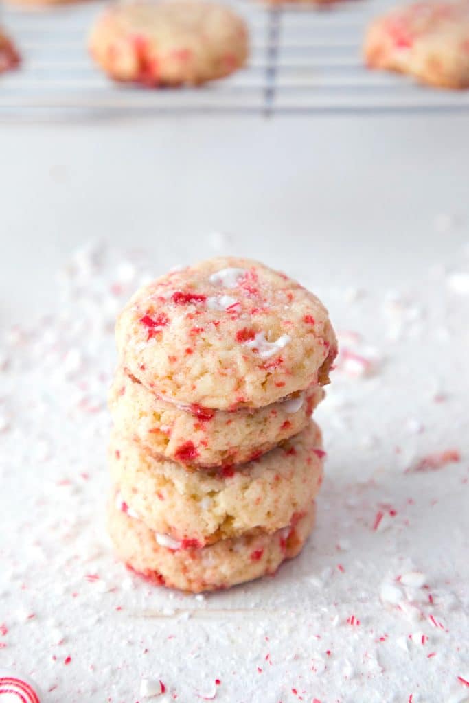 Stack of four peppermint sugar cookies on a white surface surrounded by crushed candy canes with a cookie rack and more cookies in the background