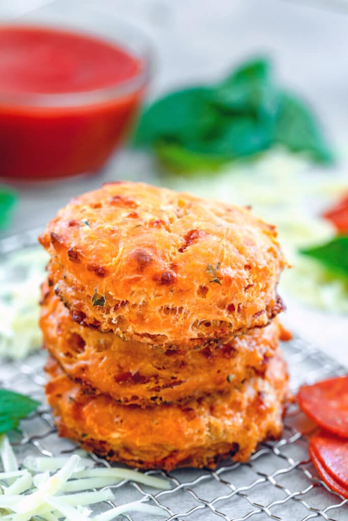 Head-on closeup view of a stack of three pepperoni pizza biscuits on a small baking rack surrounded by pepperoni, shredded cheddar cheese, and basil with small bowl of tomato sauce in background