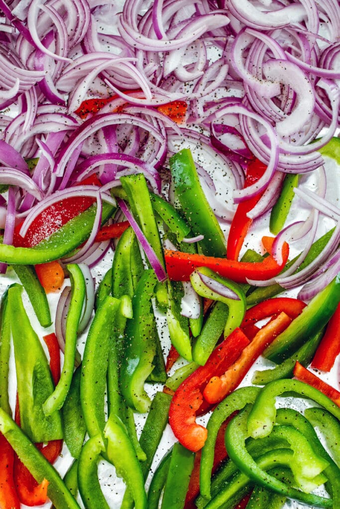 Overhead view of sliced red onion and green and red bell peppers on a baking sheet ready to go into oven