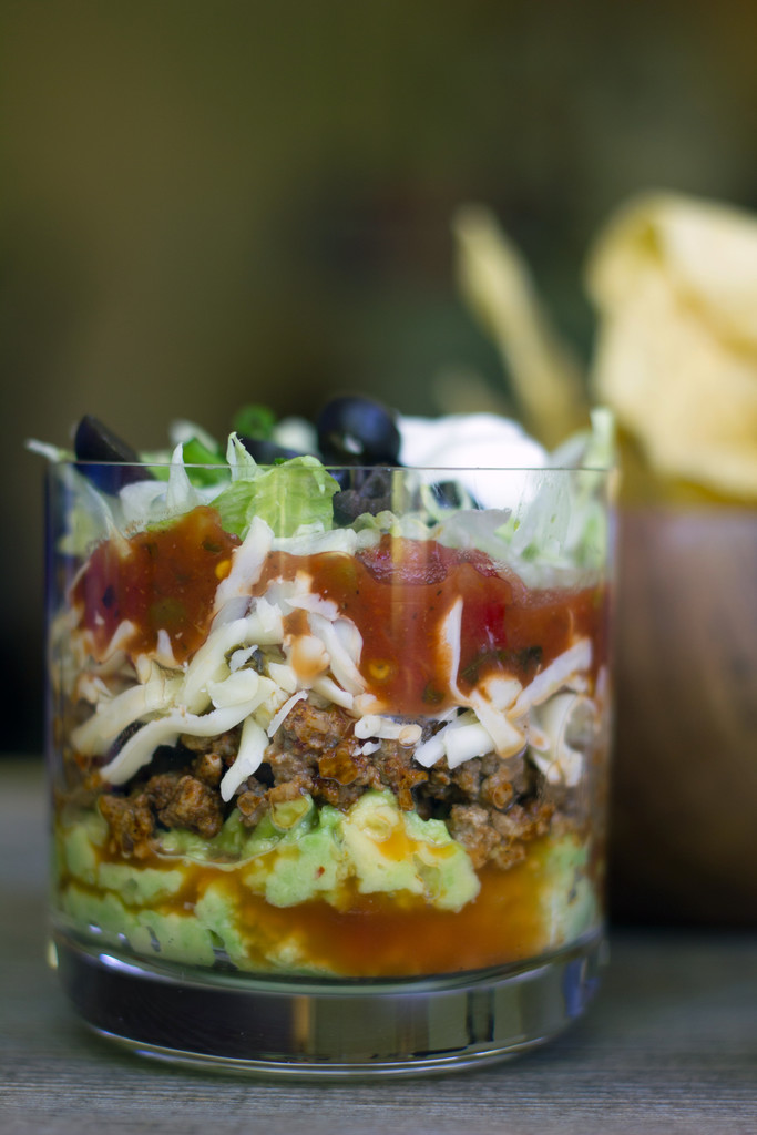 Head-on view of mini taco dips in clear glass with layers of guacamole, beef, shredded cheese, salsa, lettuce, and olives