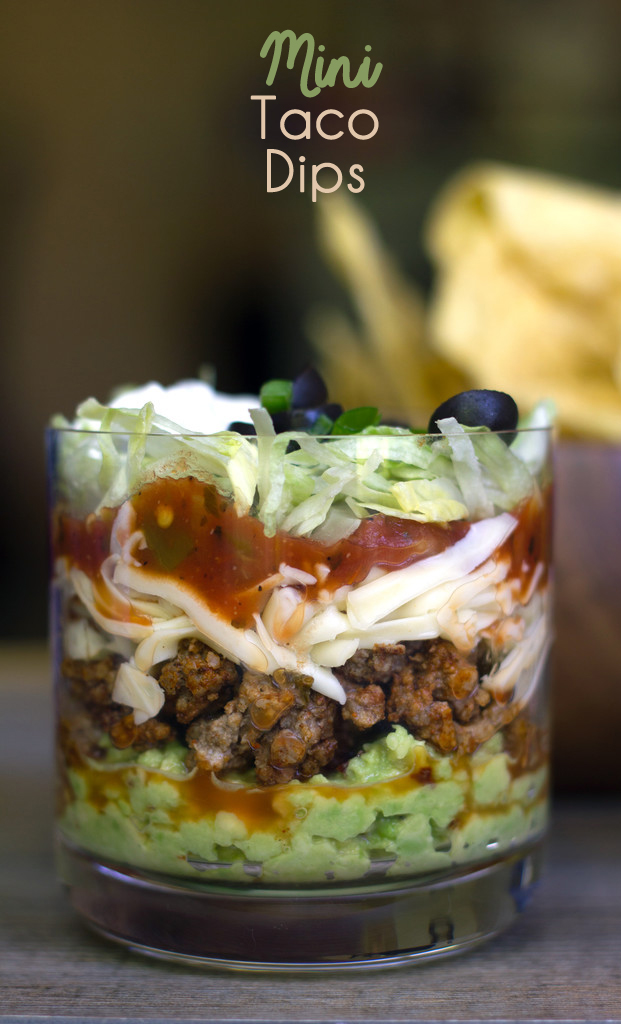 Head-on view of mini taco dips in clear glass with layers of guacamole, beef, shredded cheese, salsa, lettuce, and olives with recipe title at top