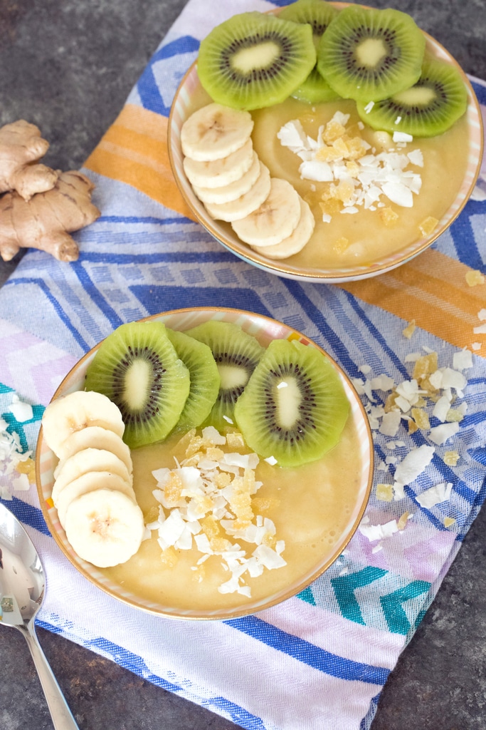 Overhead view of two pineapple banana ginger smoothie bowls with sliced bananas and kiwi and shredded coconut with coconut, ginger, and a spoon in the background