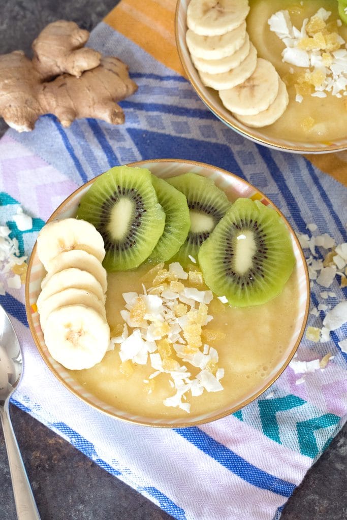 Overhead view of a pineapple banana ginger smoothie bowl topped with sliced bananas and kiwi and shredded coconut with coconut, ginger, and a spoon in the background