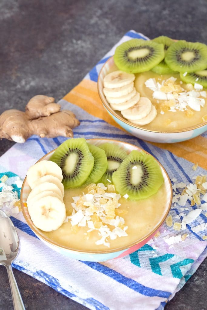 From a distance view of two pineapple banana ginger smoothie bowls with sliced bananas and kiwi and shredded coconut with a spoon, coconut, and ginger in the background