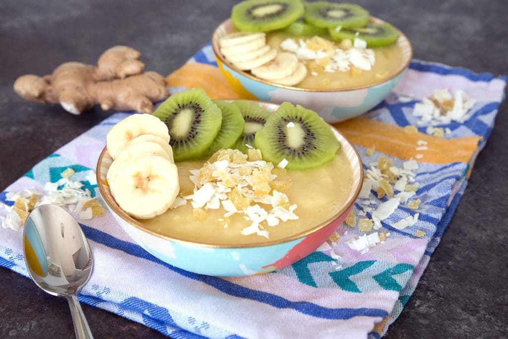 Landscape view of a pineapple banana ginger smoothie bowl with a second bowl, a spoon, coconut, and ginger in the background