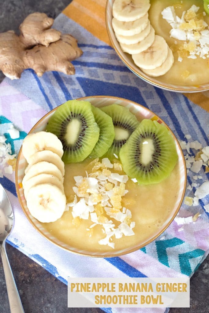 Overhead view of a pineapple banana ginger smoothie bowl topped with sliced bananas and kiwi and shredded coconut with coconut, ginger, and a spoon in the background and recipe title at the bottom