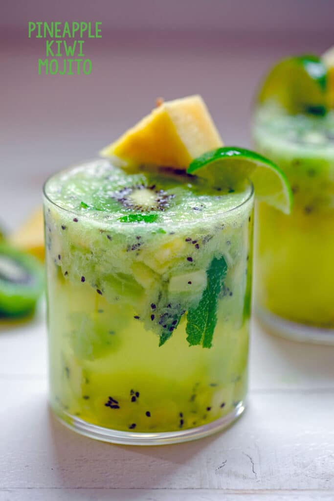 Head-on image of glass of pineapple kiwi mojito with lots of chopped pineapple and kiwi and pineapple and lime garnish with "Pineapple Kiwi Mojito" text at top.