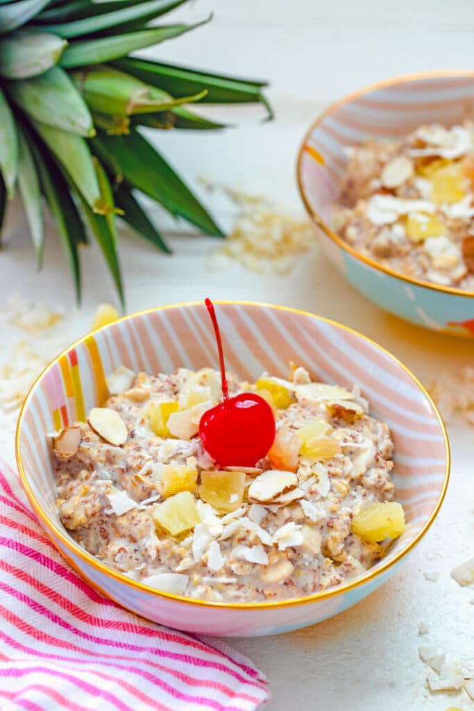 Head-on view of a bowl of piña colada bircher muesli with cherry on top and pineapple in background