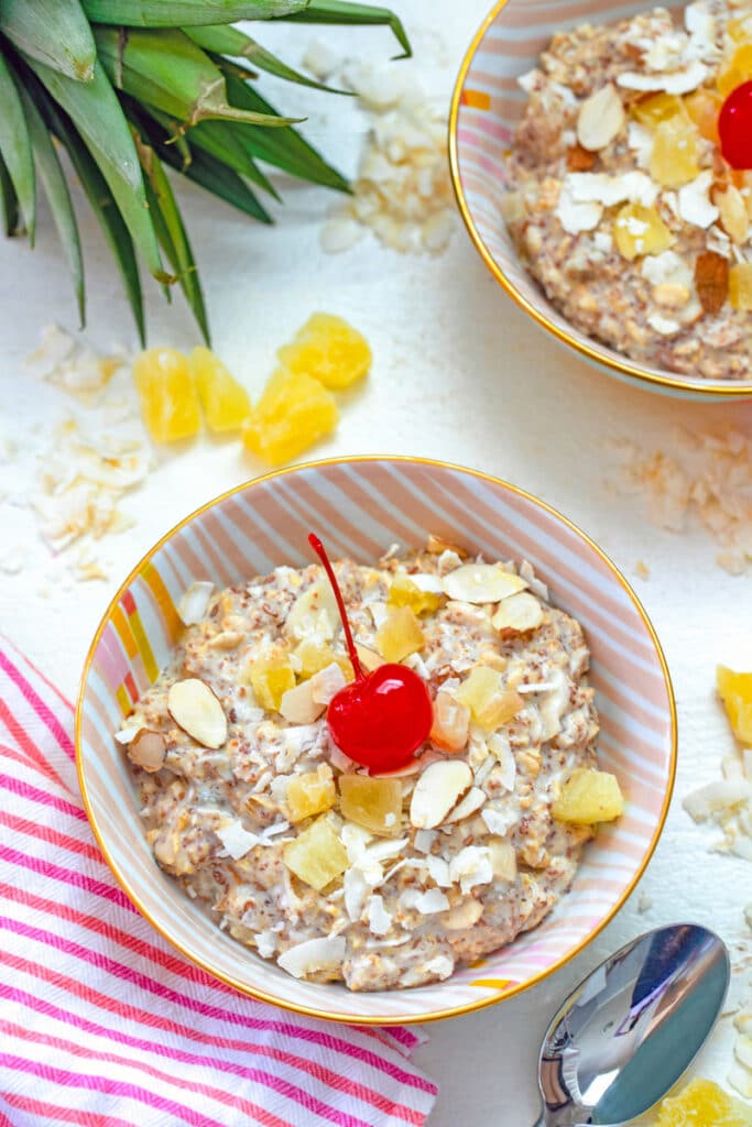 Overhead view of a bowl of piña colada bircher muesli with a cherry on top with second bowl, dried pineapple, and coconut in background