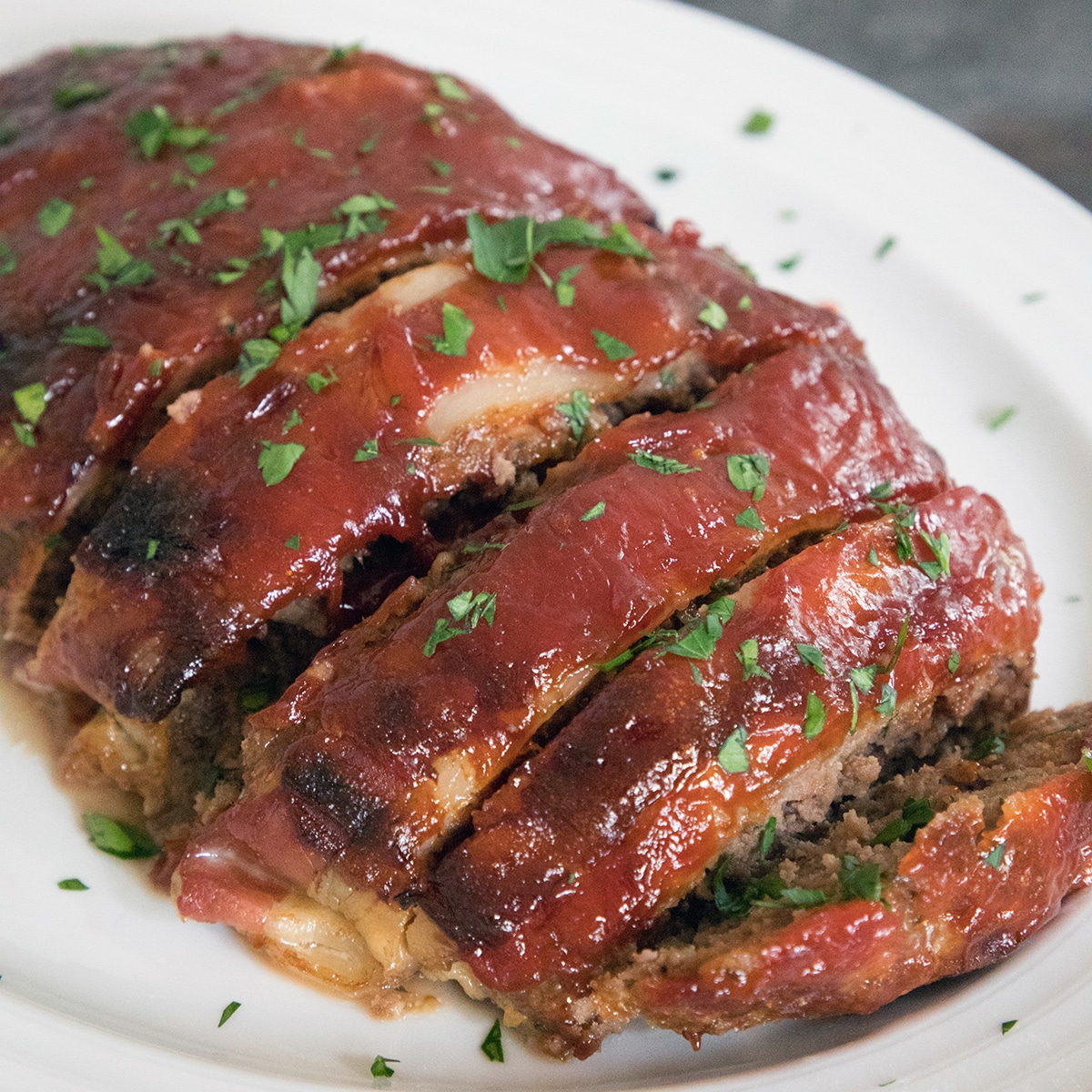 Closeup view of Bacon Meatloaf from the Pioneer Woman sliced on a white platter and covered in sauce and parsley