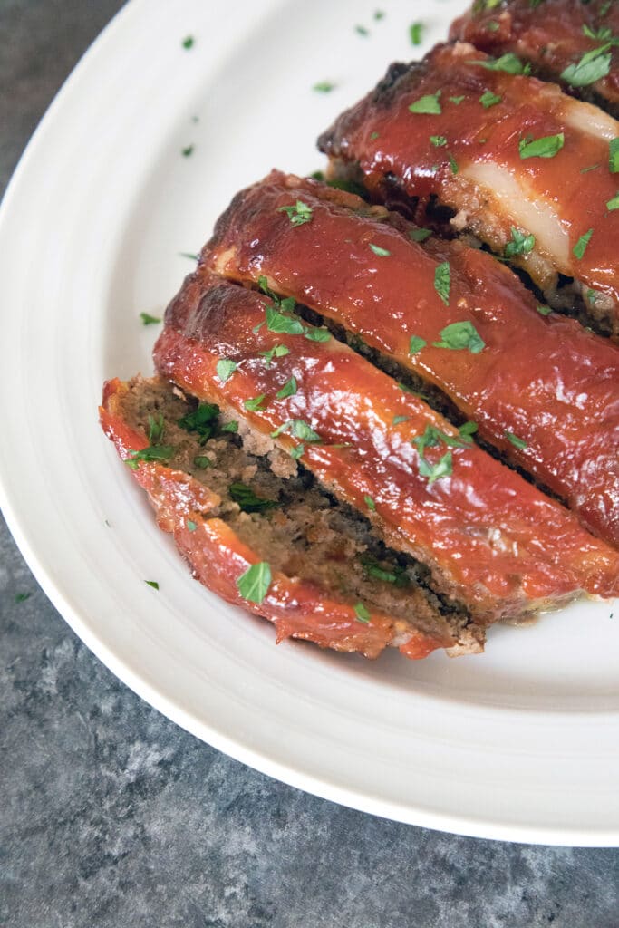 A closeup view of the slices of the Pioneer Woman's meatloaf covered in bacon, sauce, and parsley on a white platter.