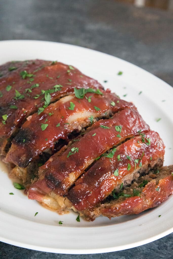 Bacon Meatloaf from the Pioneer Woman sliced on a white platter and covered in sauce and parsley.