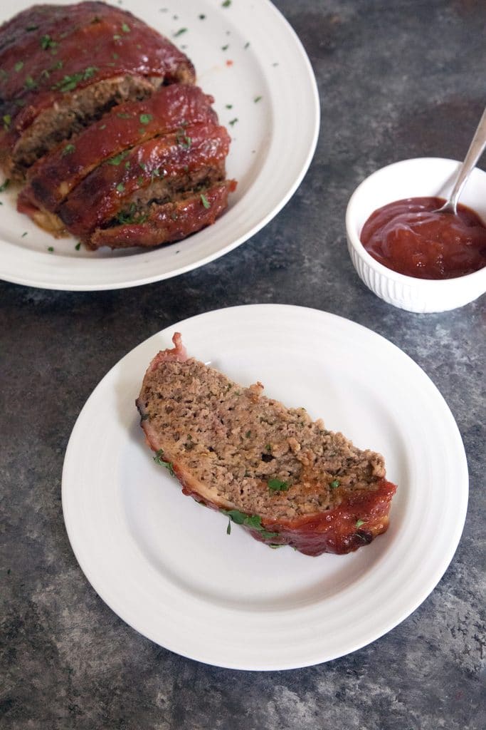 Draufsicht auf eine Scheibe Hackbraten der Pionierin auf einem weißen Teller mit einer Platte mit dem Rest des Speckhackbraten und einer Schüssel Sauce im Hintergrund's meatloaf on a white plate with a platter with the rest of the bacon meatloaf and a bowl of sauce in the background 