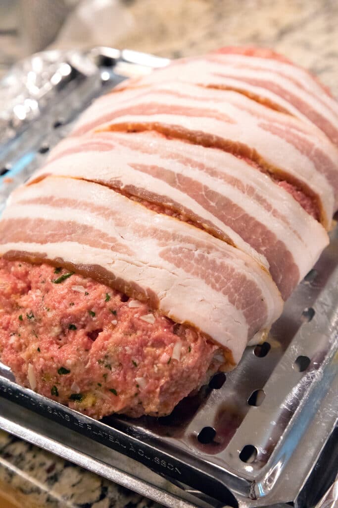 Ground beef mixture formed into meatloaf shaped and wrapped in bacon on baking pan.