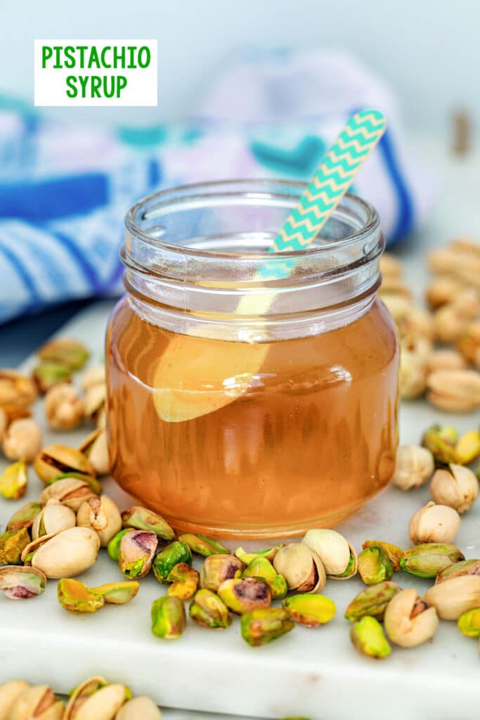 Head-on view of a small jar of pistachio syrup with a spoon in it, surrounded by lots of shelled and unshelled pistachios with recipe title at top.