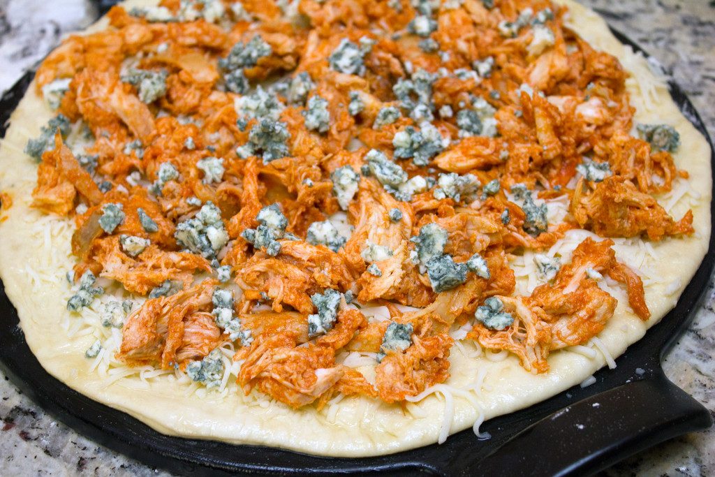 Pizza dough rolled into a round on a pizza pan and covered in mozzarella cheese, shredded buffalo chicken, and crumbled blue cheese