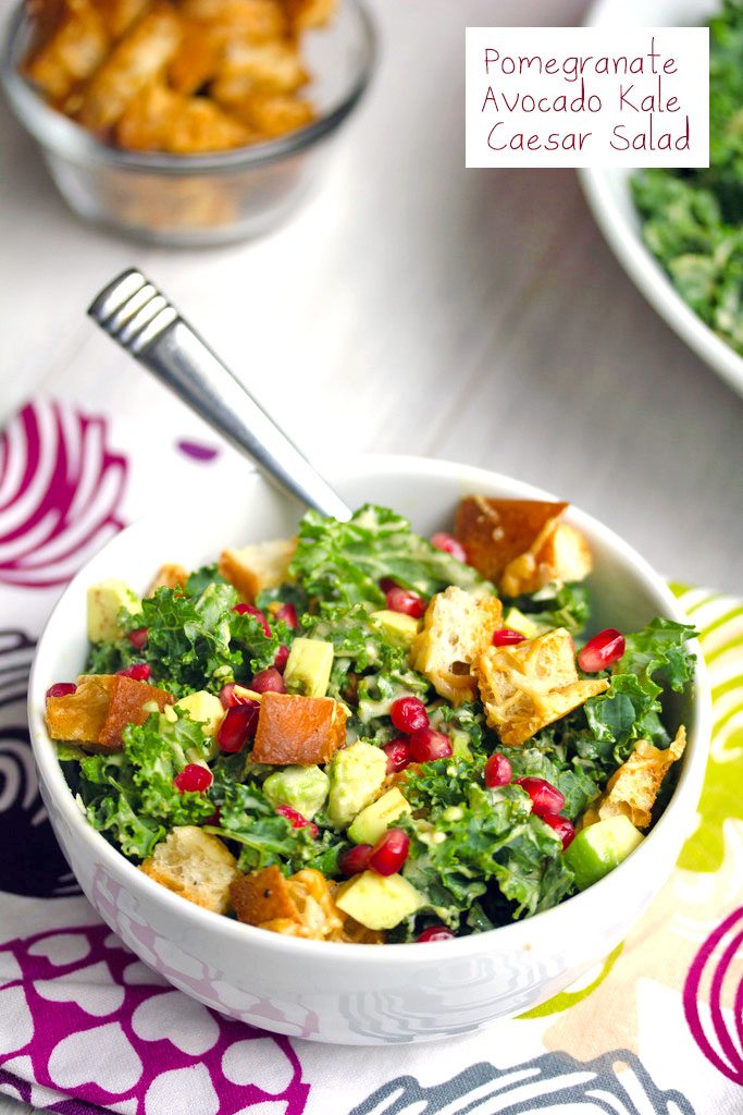 Overhead view of a pomegranate avocado kale caesar salad in a white bowl with parmesan croutons and fork with bowl of croutons in the background and recipe title at top