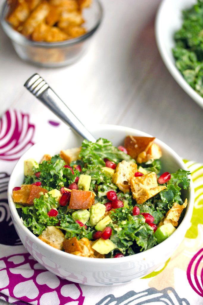 Overhead view of a pomegranate avocado kale caesar salad in a white bowl with parmesan croutons and fork with bowl of croutons in the background