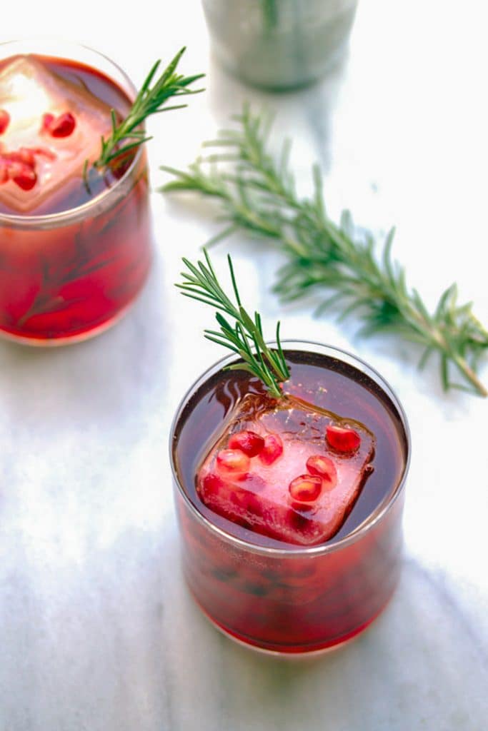 Two pomegranate and gin cocktails with rosemary in rocks glasses with pomegranate ice cubes and rosemary sprigs in background