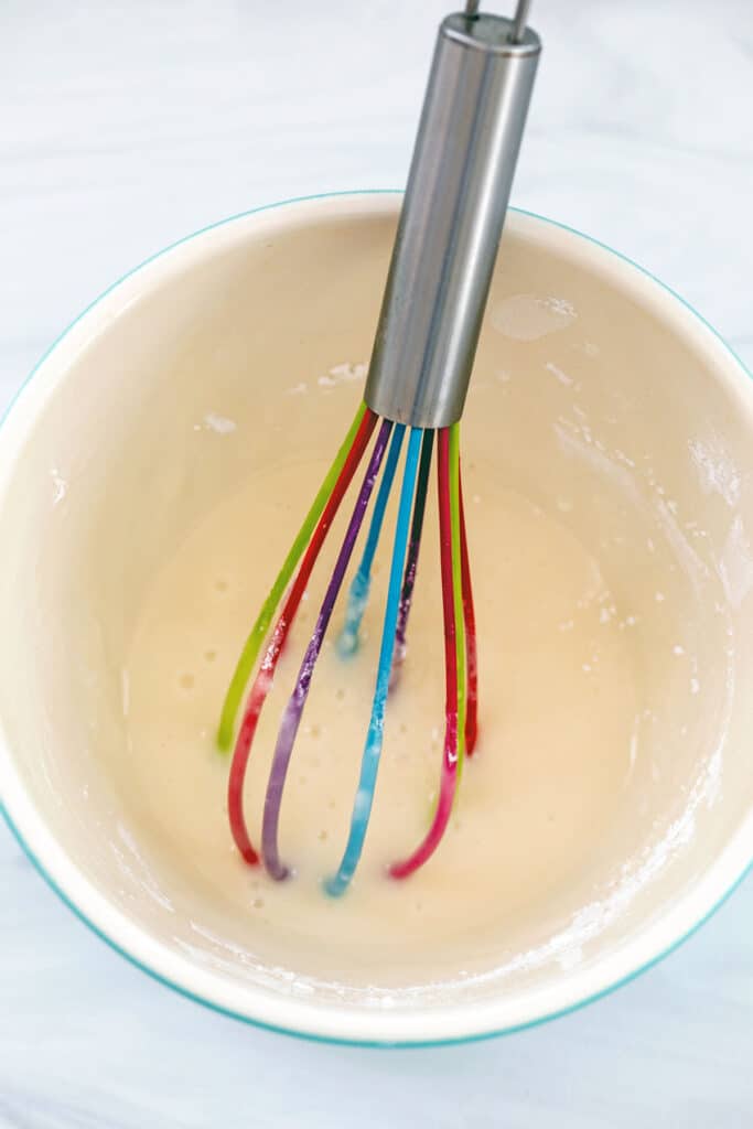Overhead view of vanilla icing in bowl with rainbow whisk.