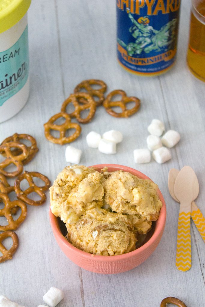Overhead view of bowl of pumpkin beer ice cream, surrounded by pretzels, mini marshmallows, wooden spoons, can of beer, glass of beer, and quart of ice cream