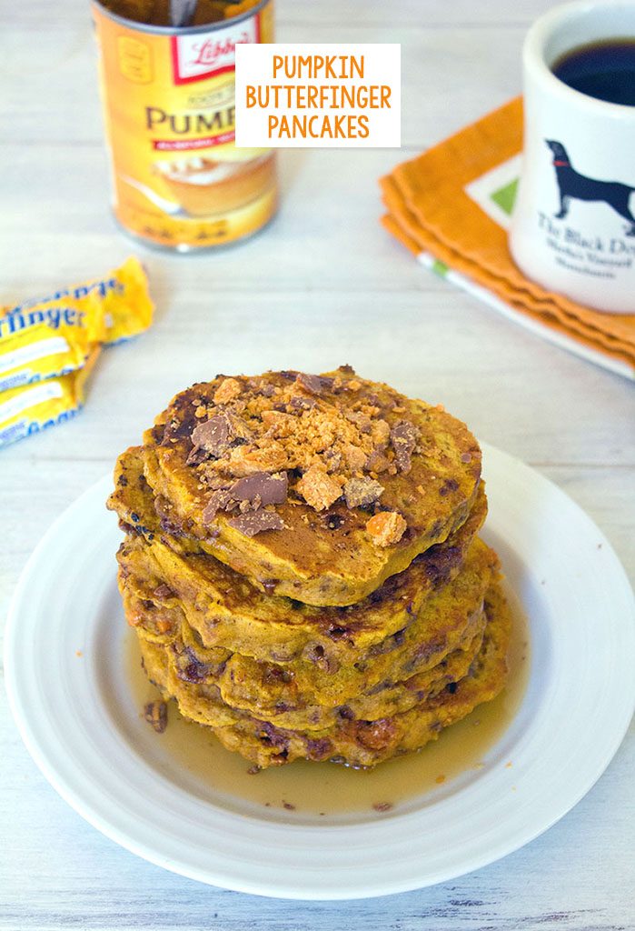 Head-on view of a stack of pumpkin butterfinger pancakes with crushed Butterfingers on top, coffee, candy, and a can of pumpkin in the background and recipe title at top