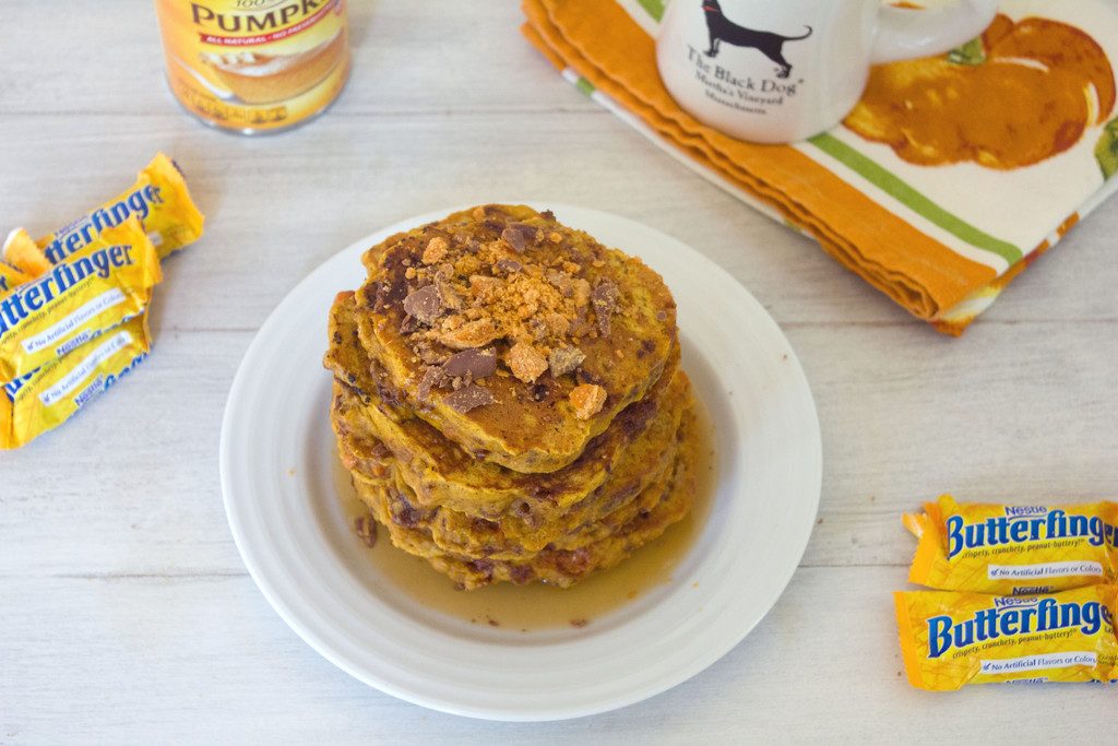 Lanscape overhead view of a stack of pumpkin butterfinger pancakes with Butterfinger candies all around and a can of pumpkin and mug of coffee in the background