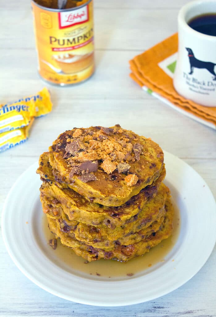 Head-on view of a stack of pumpkin butterfinger pancakes with crushed Butterfingers on top, coffee, candy, and a can of pumpkin in the background