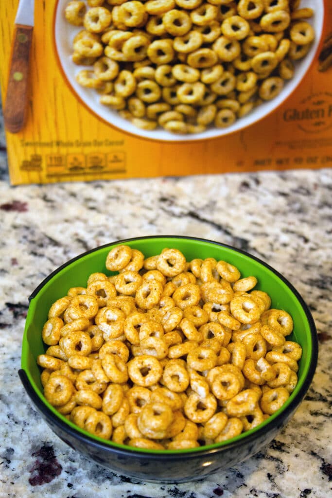 Head-on view of a measuring cup filled with Pumpkin Spice Cheerios with a box of cereal in the background