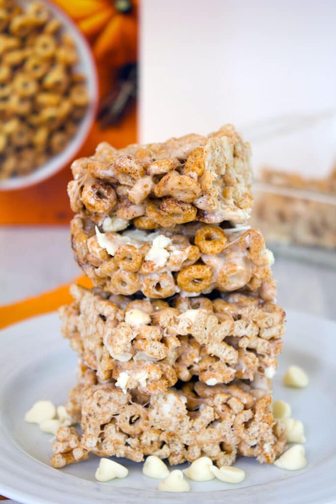 Head-on close-up view of four pumpkin Cheerios marshmallow treats on a white plate surrounded by white chocolate chips