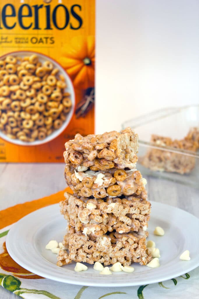 Head-on view of a stack of four pumpkin Cheerios marshmallow treats on a white plate surrounded by white chocolate chips with box of cereal and pan of bars in the background