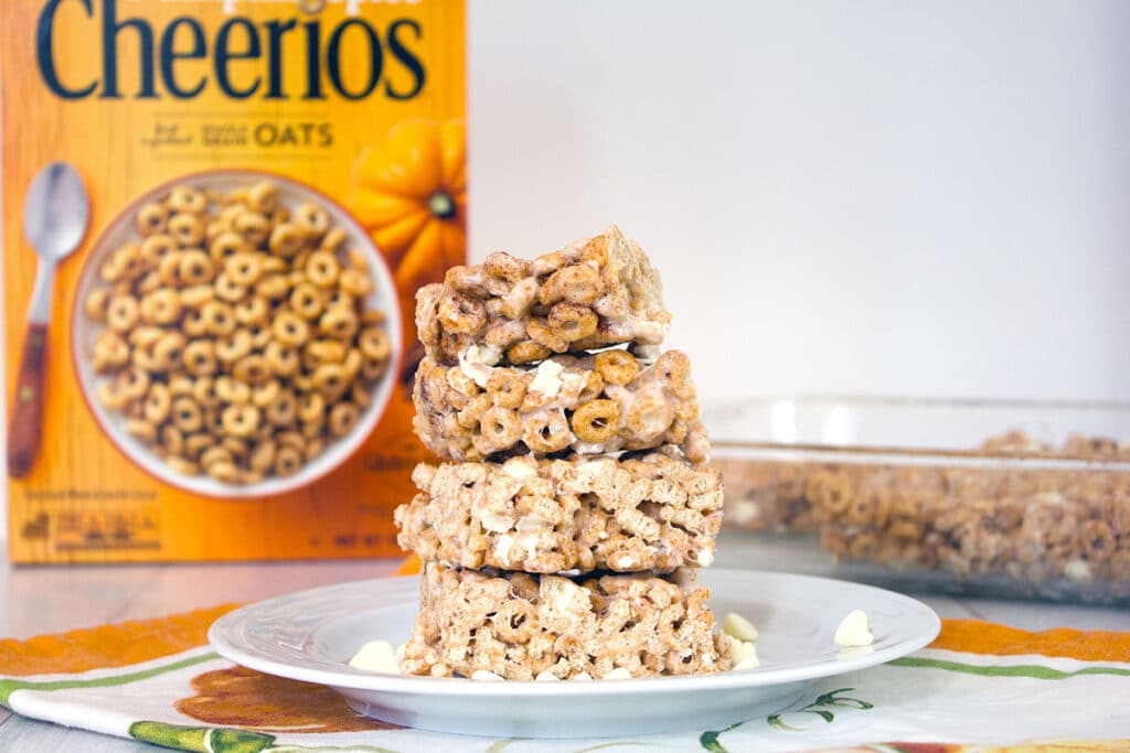 Landscape head-on view of a stack of four pumpkin Cheerios marshmallow treats on a white plate with white chocolate chips with box of cereal and more bars in pan in the background