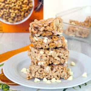 Head-on closeup view of a stack of four pumpkin Cheerios marshmallow treats on a white plate surrounded by white chocolate chips with box of cereal and pan of bars in the background