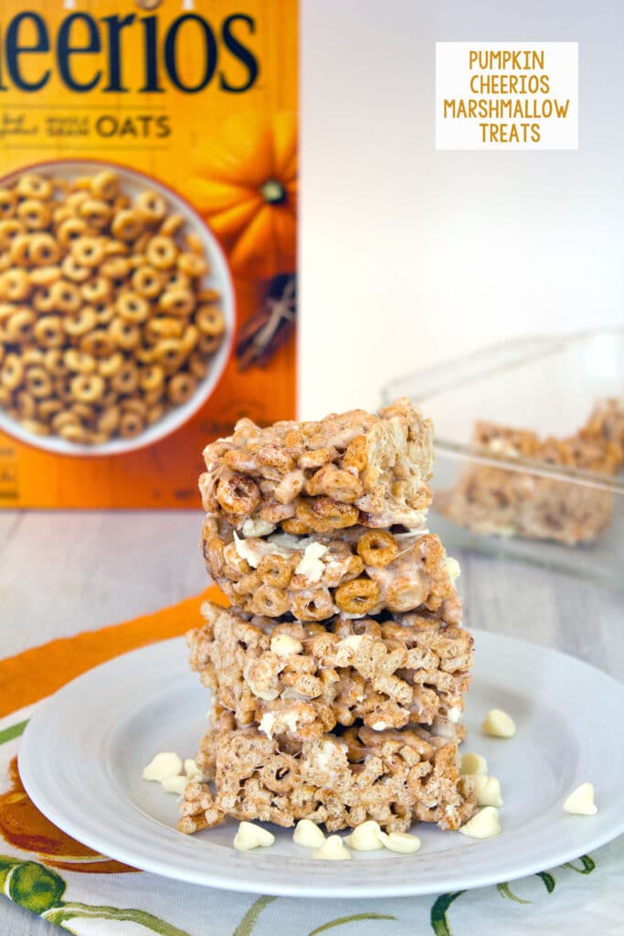 Head-on view of a stack of four pumpkin Cheerios marshmallow treats on a white plate surrounded by white chocolate chips with box of cereal and pan of bars in the background and recipe title at top