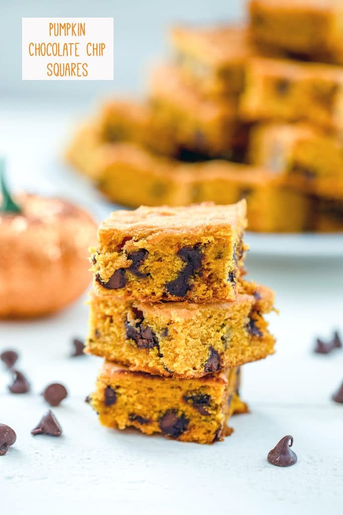 Head-on view of a stack of three pumpkin chocolate chip bars with chocolate chops scattered around and a glitter pumpkin and platter full of more bars in the background with "Pumpkin Chocolate Chips Squares" text at top