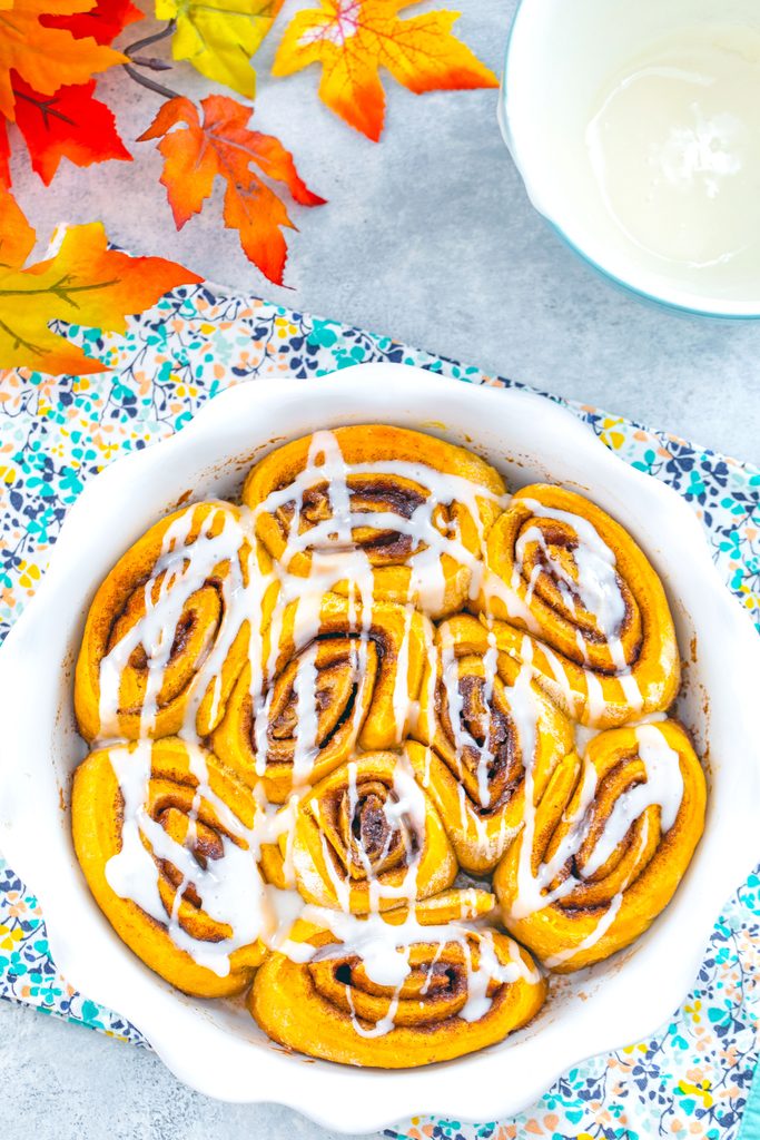 Bird's eye view of a white pie plate filled with pumpkin cinnamon rolls and drizzled in icing with fall leaves and bowl of icing in the background
