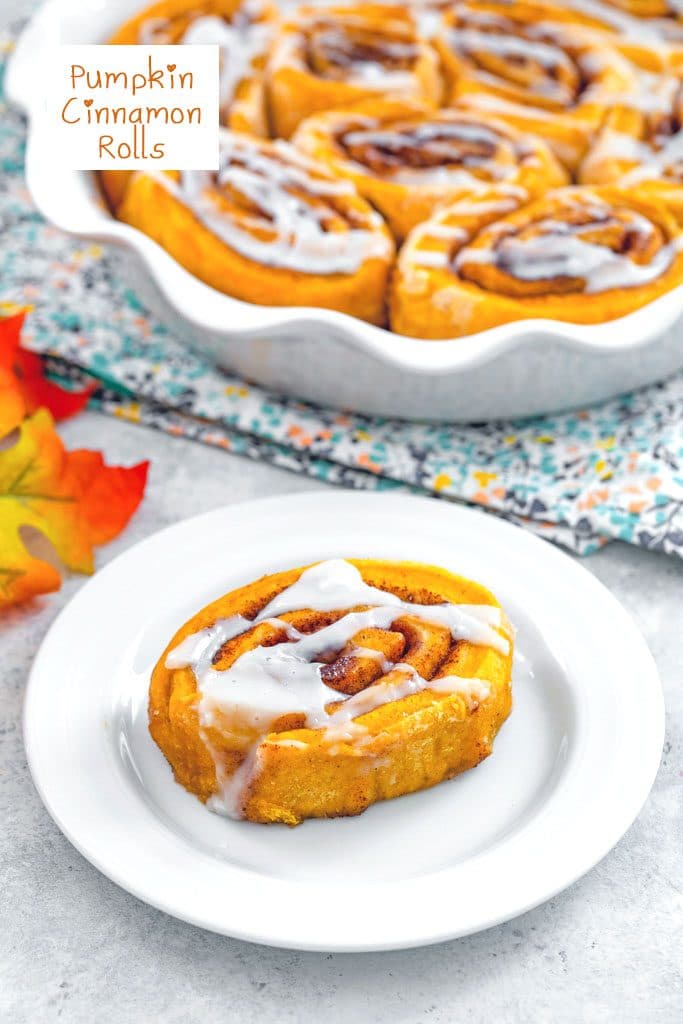 Overhead view of a pumpkin cinnamon roll with icing on a white plate with pie plate of cinnamon rolls and fall leaves in background with recipe title at top