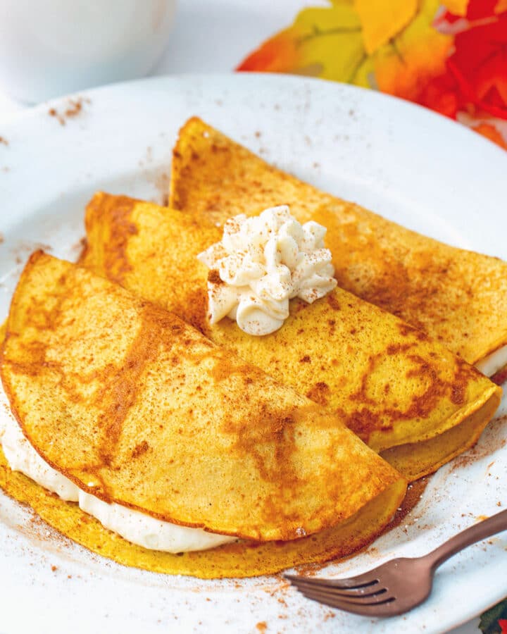 Head-on view of a plate with three pumpkin crepes filled and topped with maple cream cheese with fall leaves all around