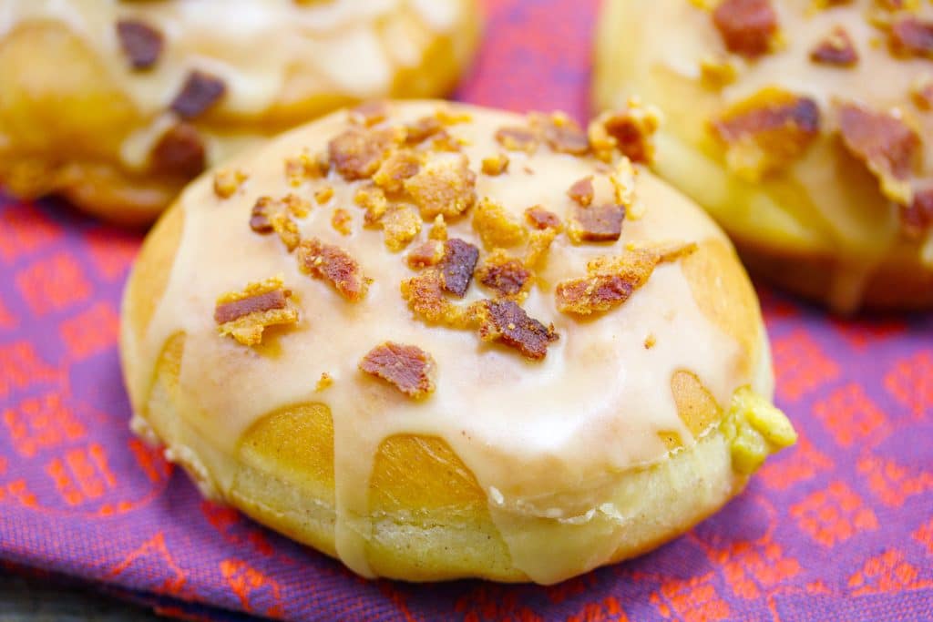 Landscape closeup of pumpkin doughnut drizzled with maple frosting and topped with crumbled bacon with pumpkin custard oozing out the front