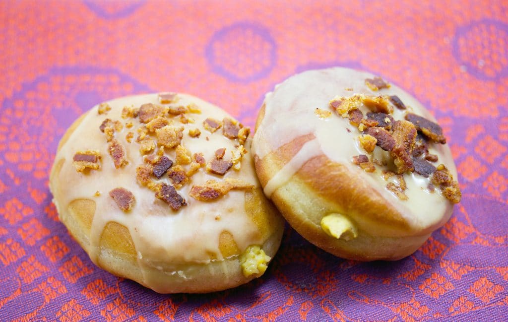 Landscape photo of two pumpkin doughnuts with maple frosting, topped with crumbled bacon with pumpkin custard oozing out the front