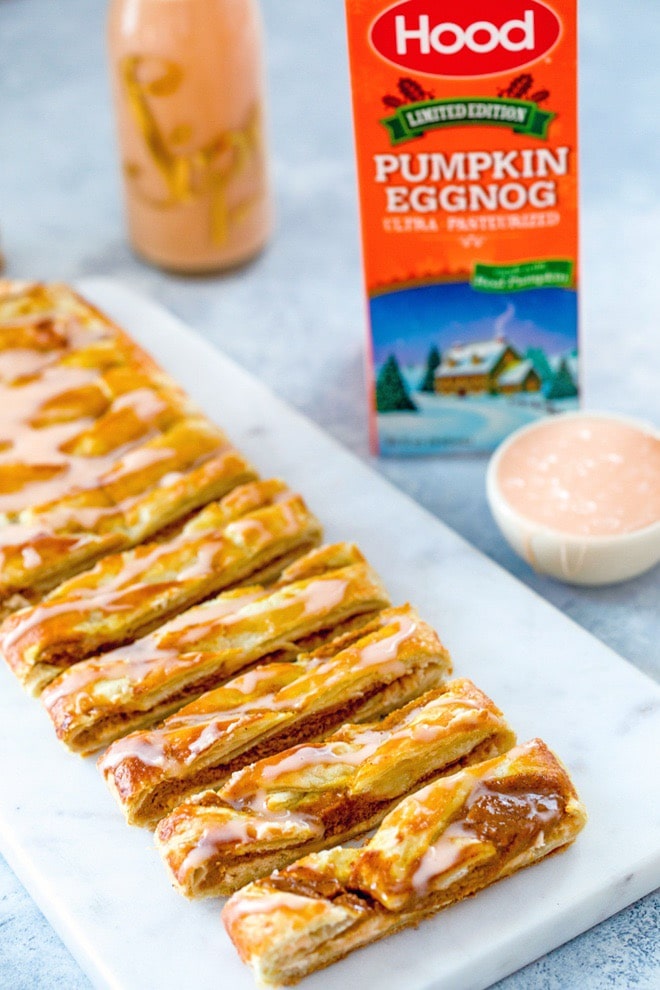 Overhead view of pumpkin eggnog danish with icing, sliced on a marble platter with carton and glass of pumpkin eggnog and bowl of pumpkin eggnog icing in the background